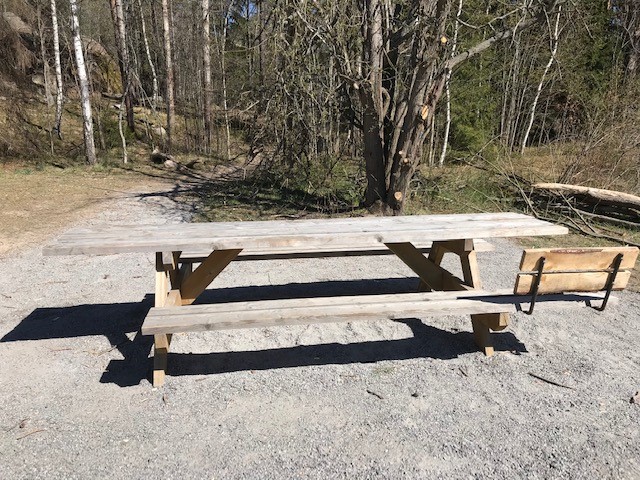 Ett bänkbord står på en yta med fint grus invid en skogsdunge.